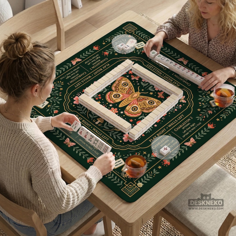 May include: A square dark green mahjong mat featuring a central moth illustration, floral patterns, and printed game instructions. The mat includes text such as Charleston 1, Charleston 2, Play Right, and Walls Left, with white tiles arranged in a square.