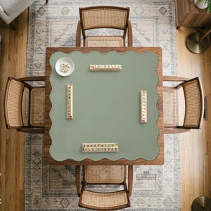 May include: A square wooden game table with a scalloped-edge sage green mat, featuring four wooden tile racks arranged around the center, a small white bowl containing game tiles, and four matching wooden chairs with woven cane backrests.