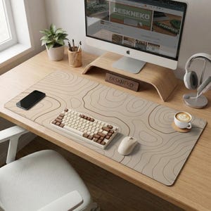 Puede incluir: Una alfombrilla de escritorio beige con un diseño topográfico, un teclado mecánico blanco con teclas marrones y un ratón blanco. Un monitor de ordenador, un soporte de monitor de madera y una taza de café también están en el escritorio. El texto "DESKNEKO" es visible.