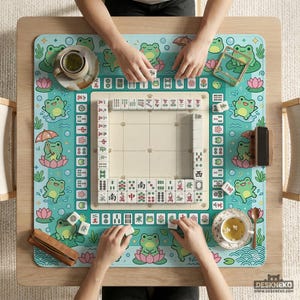 以下が含まれることがあります： 麻雀ゲームの進行状況を真上から見た様子。 ターコイズ色のマットには、カエルと蓮の花のデザインが施されています。 ゲームの駒は白地に黒文字です。 2杯の紅茶と木製のタイルホルダーも確認できます。