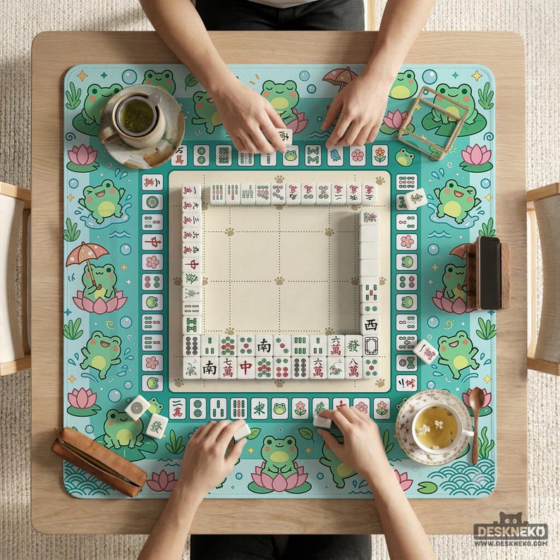 以下が含まれることがあります： 麻雀ゲームの進行状況を真上から見た様子。 ターコイズ色のマットには、カエルと蓮の花のデザインが施されています。 ゲームの駒は白地に黒文字です。 2杯の紅茶と木製のタイルホルダーも確認できます。
