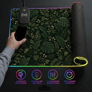K&ouml;nnte beinhalten: Eine rechteckige Schreibtischunterlage mit einem dunkelgr&uuml;nen botanischen Muster aus Monstera-Bl&auml;ttern und kleinen wei&szlig;en Bl&uuml;ten. Die Unterlage enth&auml;lt ein integriertes Qi-Ladepad, ein schwarzes Smartphone und einen RGB-LED-Lichtstreifen.