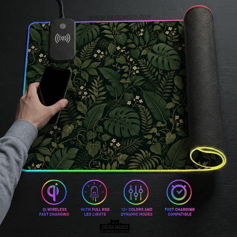 K&ouml;nnte beinhalten: Eine rechteckige Schreibtischunterlage mit einem dunkelgr&uuml;nen botanischen Muster aus Monstera-Bl&auml;ttern und kleinen wei&szlig;en Bl&uuml;ten. Die Unterlage enth&auml;lt ein integriertes Qi-Ladepad, ein schwarzes Smartphone und einen RGB-LED-Lichtstreifen.