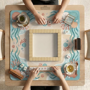 May include: A square mahjong mat featuring a light blue and beige ocean-themed pattern with coral, seashells, and pearls. The mat displays a central square of face-down mahjong tiles, a tile rack with characters, two tea cups, and a wooden phone stand.