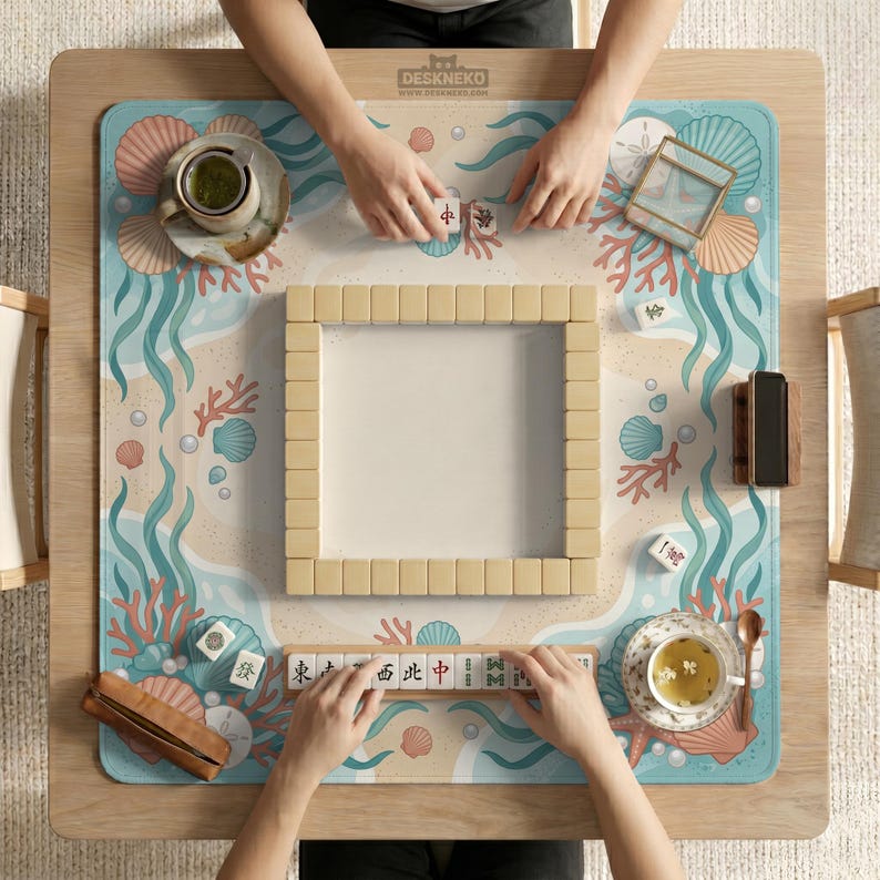 May include: A square mahjong mat featuring a light blue and beige ocean-themed pattern with coral, seashells, and pearls. The mat displays a central square of face-down mahjong tiles, a tile rack with characters, two tea cups, and a wooden phone stand.