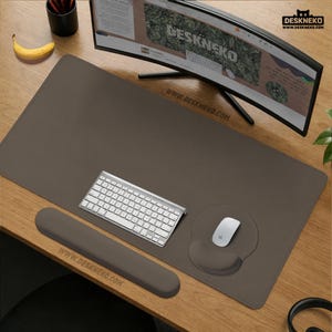 May include: A brown desk mat with a matching wrist rest and mouse pad. A white keyboard and mouse are on the mat. The monitor displays the text "DESKNEKO" and the website address "www.deskneko.com".