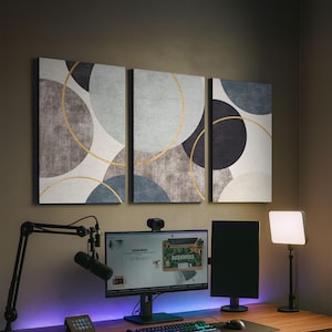 Puede incluir: Tríptico de arte mural abstracto con círculos superpuestos en gris, azul y negro, resaltados por líneas doradas. La obra de arte está montada sobre un escritorio con equipo informático, incluyendo un monitor, micrófono y teclado.