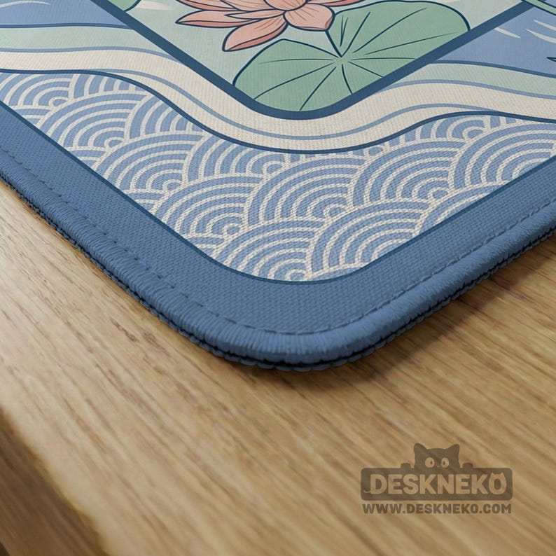 May include: A rectangular desk mat with a tranquil design of a lotus flower, water, and wave patterns in blue, green, and pink. The mat has a blue border and the text "DESKNEKO" and "WWW.DESKNEKO.COM".