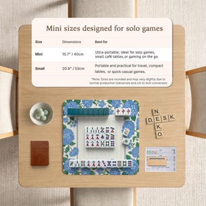 May include: A mini Mahjong set on a wooden table. The set includes tiles, a small brown notebook, and a bowl with small green stones. The Mahjong mat has a blue floral design. The text on the table reads "Mini sizes designed for solo games."