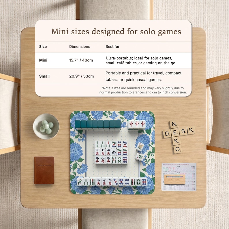 May include: A mini Mahjong set on a wooden table. The set includes tiles, a small brown notebook, and a bowl with small green stones. The Mahjong mat has a blue floral design. The text on the table reads "Mini sizes designed for solo games."