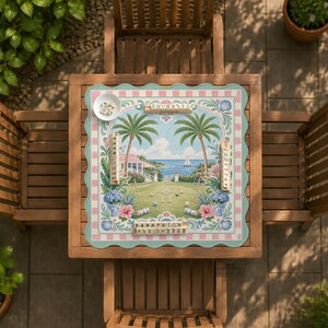 May include: A square croquet game board with a coastal scene, featuring palm trees, a house, and a sailboat. The board is set on a wooden table with two chairs. The board has a pink and blue checkered border and a decorative edge.