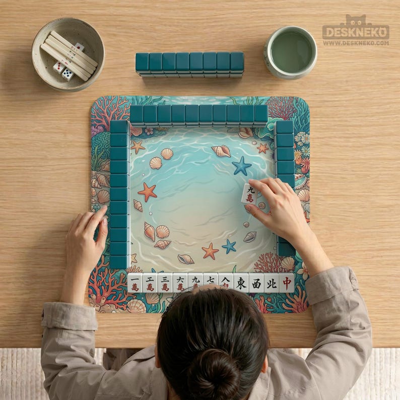 Pode incluir: Uma pessoa jogando Mahjong em uma mesa de madeira. O tabuleiro apresenta uma cena de praia com conchas e estrelas do mar. As pe&ccedil;as turquesas est&atilde;o dispostas ao redor do tabuleiro. Outras pe&ccedil;as do jogo incluem dados e uma tigela pequena.