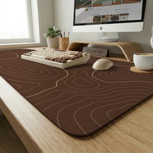 May include: A brown desk mat with a topographic design in white lines. A white keyboard, mouse, and a computer monitor are on the desk. The desk mat is rectangular and covers a large portion of the desk surface.