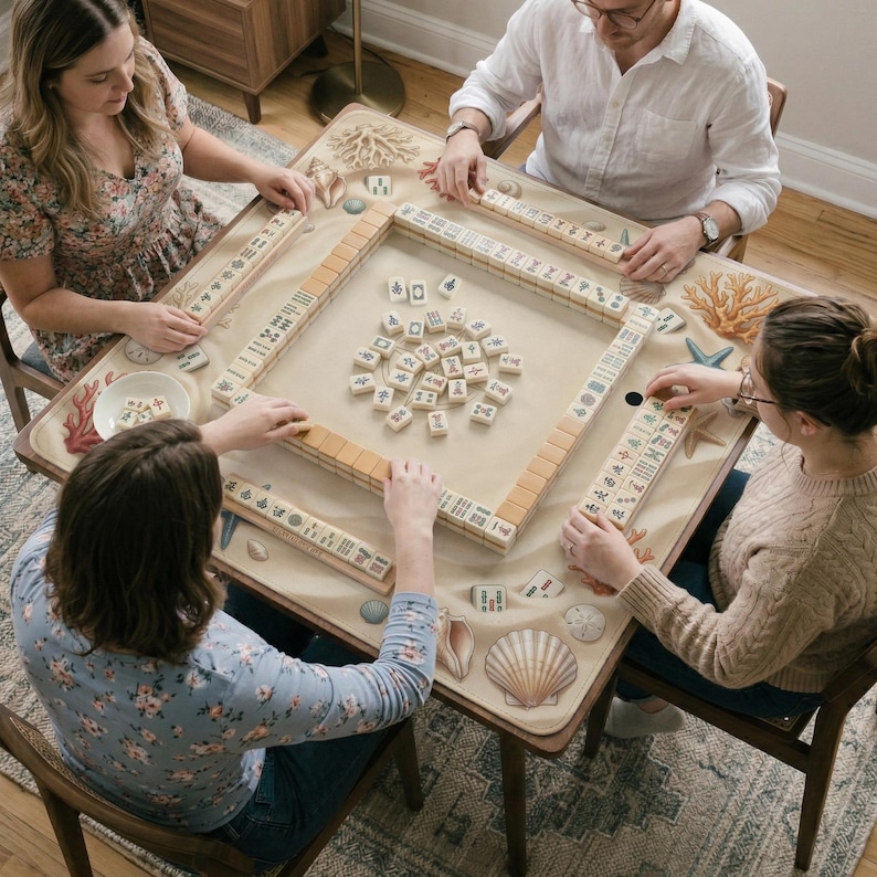以下が含まれることがあります： 麻雀をしている人々のグループ。ベージュのゲームテーブルは、貝殻とサンゴの沿岸デザインが特徴です。プレイヤーは、黒いシンボルが付いたタン色のタイルを配置しています。テーブルは木製の椅子に囲まれています。