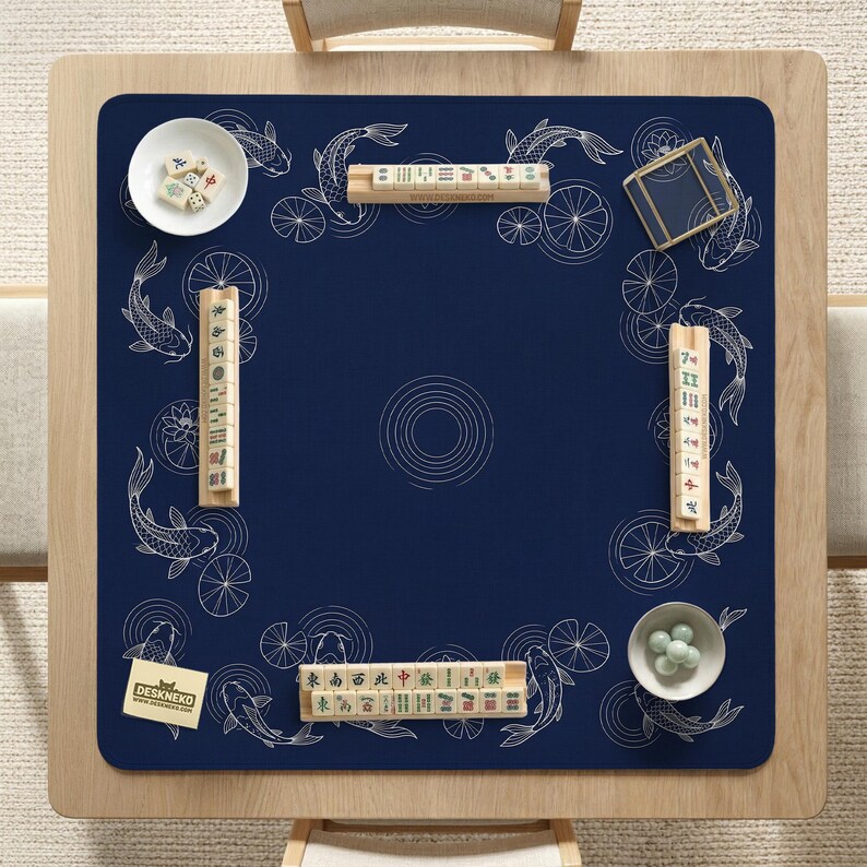 Puede incluir: Una mesa de mahjong cuadrada de madera con una alfombrilla azul marino con dise&ntilde;os de peces koi blancos y flores de loto. La mesa est&aacute; puesta con fichas de mahjong, dados y cuencos peque&ntilde;os. Se ve una tarjeta peque&ntilde;a con el texto "DESKNEKO".