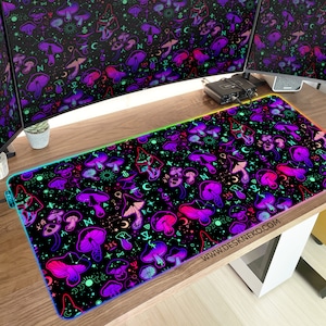 May include: A rectangular black desk mat with a vibrant pattern of purple and pink mushrooms, moons, and stars. The mat has a glowing rainbow edge and the website address www.deskneko.com printed on it. The mat is on a wooden desk.