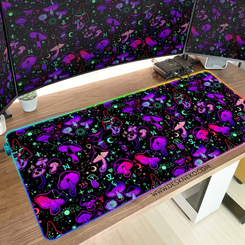 May include: A rectangular black desk mat with a vibrant pattern of purple and pink mushrooms, moons, and stars. The mat has a glowing rainbow edge and the website address www.deskneko.com printed on it. The mat is on a wooden desk.