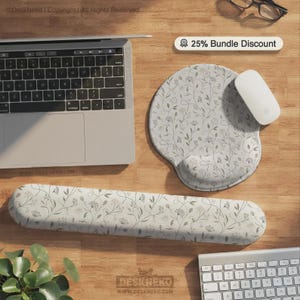 Pu&ograve; includere: Un tappetino per mouse grigio chiaro con poggiapolsi integrato e un lungo poggiapolsi per tastiera abbinato, entrambi caratterizzati da un delicato motivo floreale botanico grigio scuro. Un mouse bianco &egrave; posizionato sul tappetino accanto a un laptop.