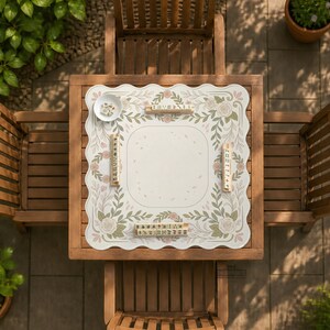 May include: A square scalloped-edge game mat featuring a cream-colored center with a floral border of sage green leaves and soft pink roses. Four wooden tile racks are placed along the edges, with a small white bowl containing game tiles.
