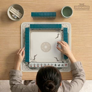 May include: A person playing Mahjong. The game set includes teal tiles, dice, and a bowl. The playing surface is white with a crane design and Chinese characters. The game is set on a light wood table.