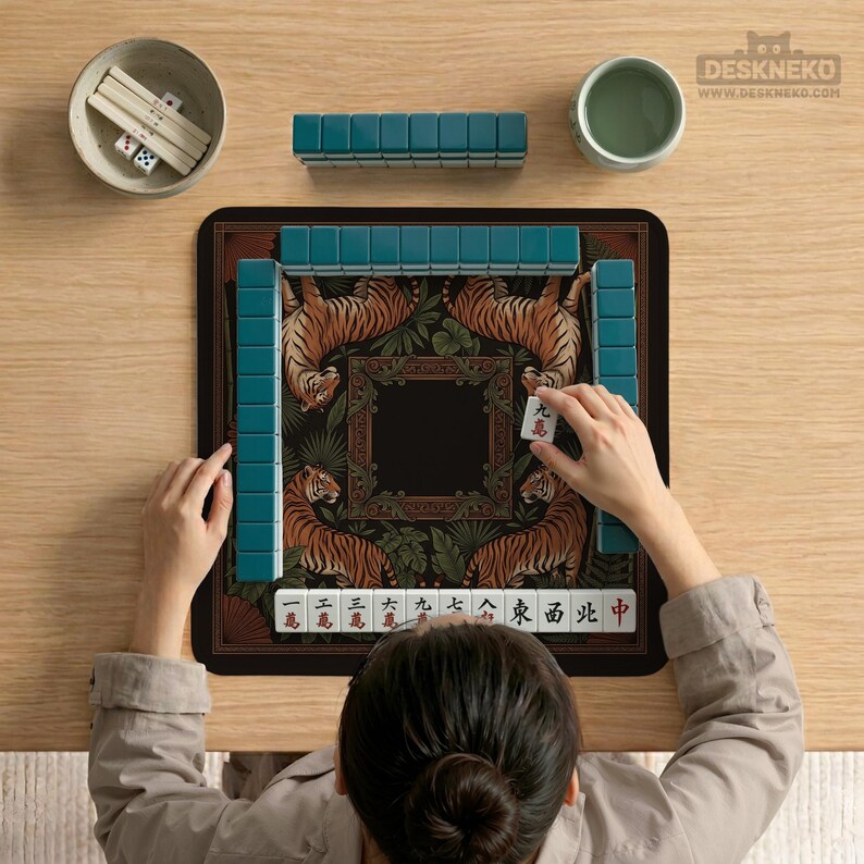 May include: A person playing Mahjong on a wooden table. The game set includes teal tiles, a black mat with tiger illustrations, a small bowl with dice, and a cup of tea. The mat features a decorative border and a central black square.