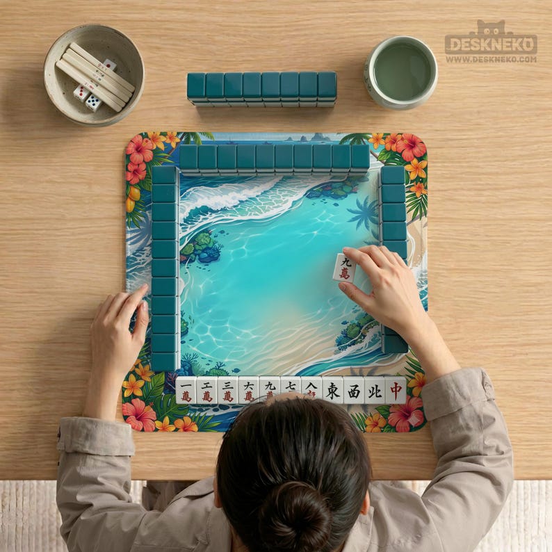 May include: A square mahjong mat featuring a tropical beach scene with turquoise water, white sand, and hibiscus flowers. The mat is surrounded by dark teal mahjong tiles, with a row of tiles at the bottom displaying red and black Chinese characters.