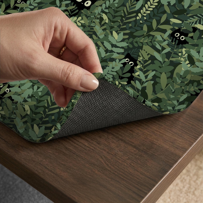 May include: A decorative mat featuring a repeating pattern of black cats peeking through green foliage. The mat's surface displays a dense arrangement of leaves in various shades of green, with the cats' faces adding a playful element. The underside is a dark gray, textured material.