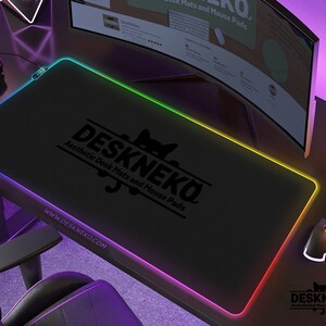 Op de afbeelding: Zwarte rechthoekige bureaumat met een gloeiende veelkleurige rand. De mat heeft de tekst "DESKNEKO Aesthetic Desk Mats and Mouse Pads" en het websiteadres www.DESKNEKO.COM. Een gamingmuis en controller zijn zichtbaar.