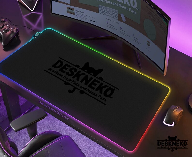 Op de afbeelding: Zwarte rechthoekige bureaumat met een gloeiende veelkleurige rand. De mat heeft de tekst "DESKNEKO Aesthetic Desk Mats and Mouse Pads" en het websiteadres www.DESKNEKO.COM. Een gamingmuis en controller zijn zichtbaar.