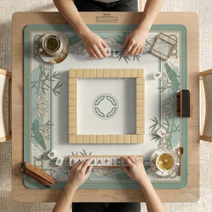 May include: A top-down view of a mahjong game in progress. The game is set on a wooden table with a floral and bamboo-themed mat. Beige tiles are arranged in a square, with players' hands visible. The text "DESKNEKO" is visible on the table.