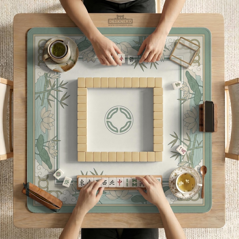 May include: A top-down view of a mahjong game in progress. The game is set on a wooden table with a floral and bamboo-themed mat. Beige tiles are arranged in a square, with players' hands visible. The text "DESKNEKO" is visible on the table.