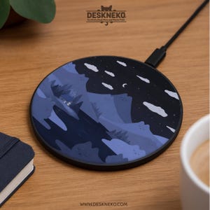 Puede incluir: Un cargador inalámbrico redondo negro con un diseño de montaña y cielo nocturno. El cargador presenta una combinación de colores azul oscuro y negro con acentos de nubes y luna blancos. Tiene un cable negro adjunto. La marca "Deskneko" está en la parte superior.