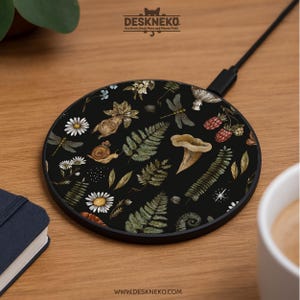 Puede incluir: Un cargador inalámbrico redondo negro con un diseño de temática natural. El cargador presenta ilustraciones de libélulas, setas, helechos, margaritas y bayas. El texto "DESKNEKO" y "www.deskneko.com" son visibles.