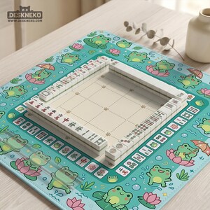 以下が含まれることがあります： ベージュ色のゲーム盤に配置された白い牌を使った麻雀セット。セットは、漫画の蛙、蓮の葉、花で飾られたターコイズ色のマットの上に置かれています。マットには「DESKNEKO」と「www.deskneko.com」の文字が入った縁があります。