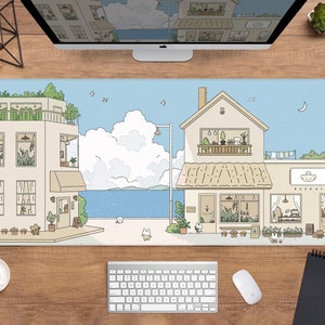 May include: A large rectangular mousepad with an illustrated town scene, including a bakery, buildings, and a blue sky. The design features cartoon animals, plants, and a seaside view. The mousepad is on a wooden desk with a computer and accessories.