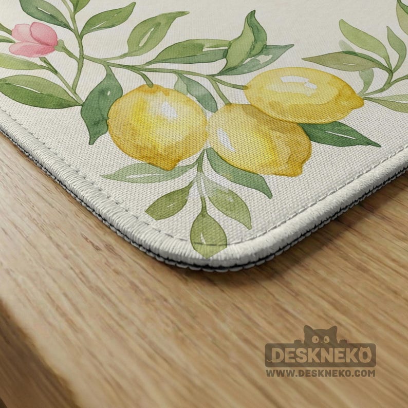 May include: A rectangular desk mat featuring a watercolor design of lemons, green leaves, and a pink flower. The mat has a cream-colored background and a black stitched border. The Deskneko logo is visible in the bottom right corner.