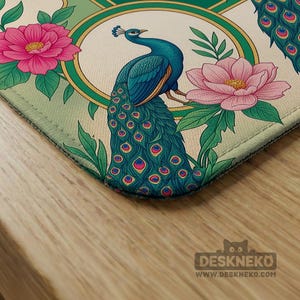 May include: A rectangular desk mat featuring a vibrant illustration of a blue peacock with a long, detailed tail, surrounded by large pink flowers and green leaves. The design is framed by a circular gold border against a light cream background.