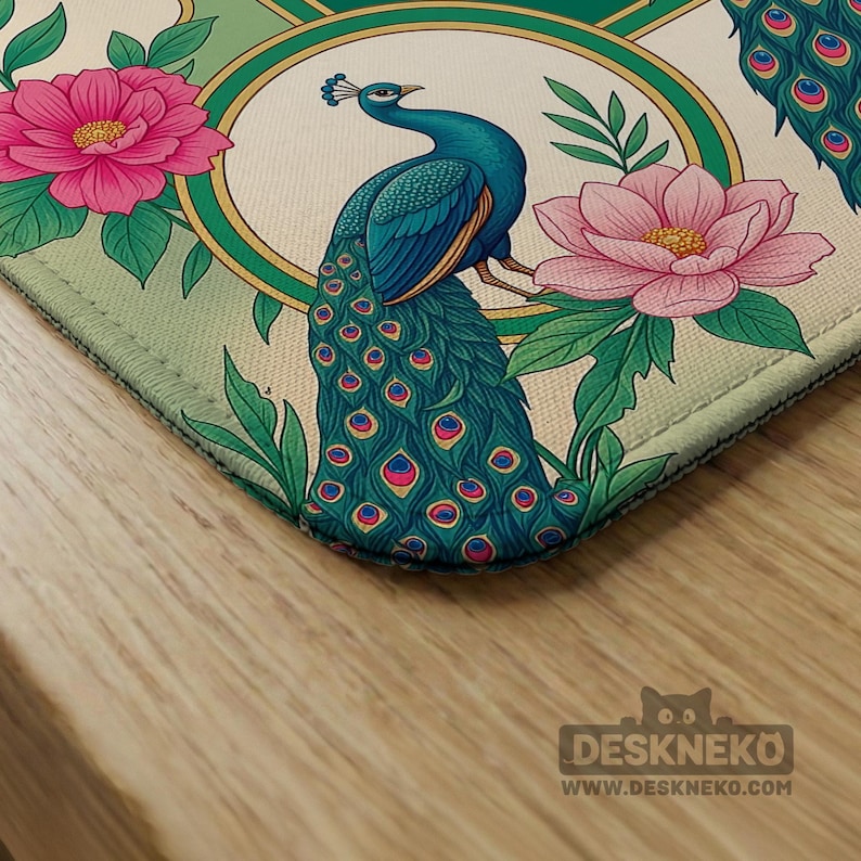 May include: A rectangular desk mat featuring a vibrant illustration of a blue peacock with a long, detailed tail, surrounded by large pink flowers and green leaves. The design is framed by a circular gold border against a light cream background.