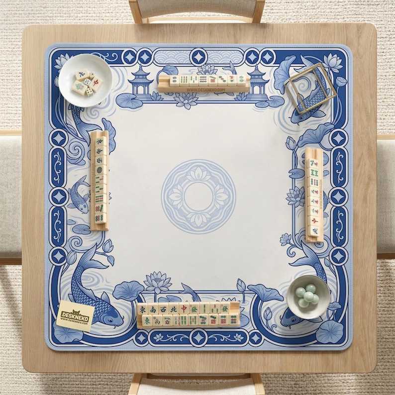 May include: A square mahjong mat featuring a blue and white pattern of koi fish, lotus flowers, and pagodas. The mat includes four wooden tile racks, two white bowls containing mahjong tiles and green marbles, and a small card labeled DESKNEKO.