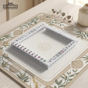 May include: A Mahjong set with white tiles and red and green markings is arranged on a beige mat with floral and seashell designs. The mat is on a light-colored wooden surface. The logo "DESKNEKO" is visible in the top left corner.