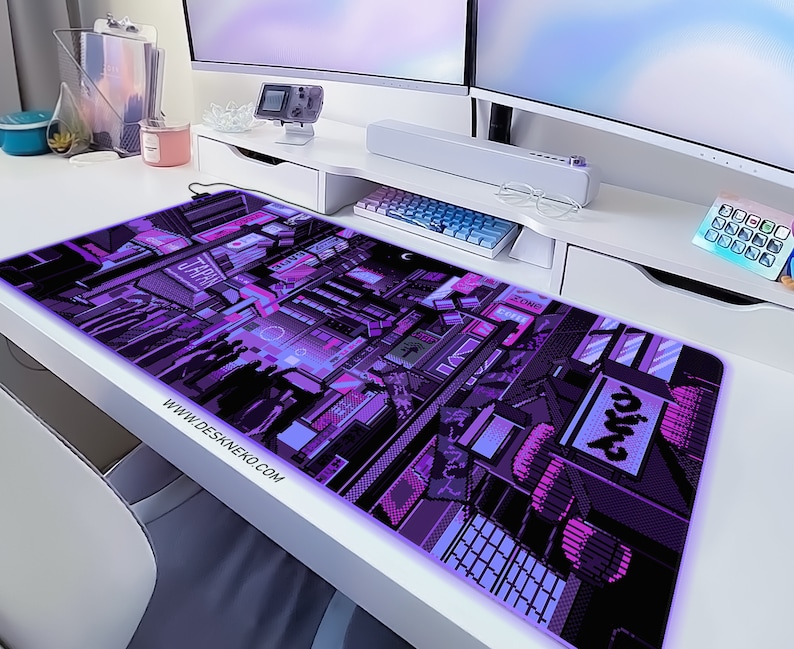 K&ouml;nnte beinhalten: Ein rechteckiges, lila-schwarzes Pixel-Art-Mauspad mit einem Stadtlandschaftsdesign. Das Mauspads hat einen leuchtenden Rand und der Text "WWW.DESKNEKO.COM" ist unten links aufgedruckt.