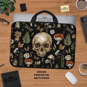 Op de afbeelding: Zwarte laptoptas met een schedel, paddenstoelen, vlinders en botanische elementen. Het ontwerp is aan beide zijden bedrukt. De tas heeft een zwart handvat en ligt op een houten oppervlak.