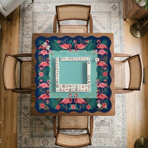 May include: A square wooden table with a blue and teal flamingo-themed Mahjong mat. The mat features pink flamingos, tropical flowers, and green leaves. The table is surrounded by four rattan chairs, and a set of Mahjong tiles is in the center.