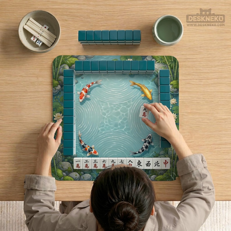 May include: A Mahjong set featuring a square play mat with a pond illustration containing four koi fish. The set includes teal tiles, a bowl with white dice and sticks, and a tile with the character 九萬 held by a hand. The mat border shows bamboo.