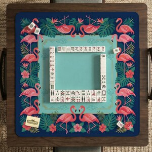 May include: A blue Mahjong game mat with a flamingo and tropical leaf design. The mat has a turquoise center and is surrounded by pink flamingos, flowers, and green leaves. The mat has a rectangular shape and is surrounded by a wooden frame.