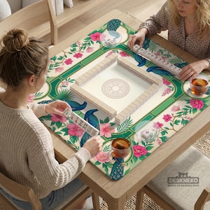 May include: A square mahjong mat featuring a cream background with a green border, decorated with pink hibiscus flowers, green foliage, and blue peacock illustrations. The mat includes a central square tile rack and individual tile sets.