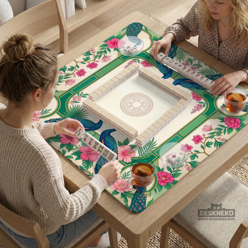 May include: A square mahjong mat featuring a cream background with a green border, decorated with pink hibiscus flowers, green foliage, and blue peacock illustrations. The mat includes a central square tile rack and individual tile sets.
