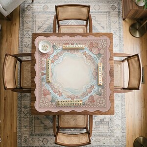 May include: Overhead view of a wooden table with four chairs. The table features a pink mat with a beach-themed design, including seashells, starfish, and a compass rose. A small bowl with game pieces sits on the mat. The chairs have woven backs and seats.