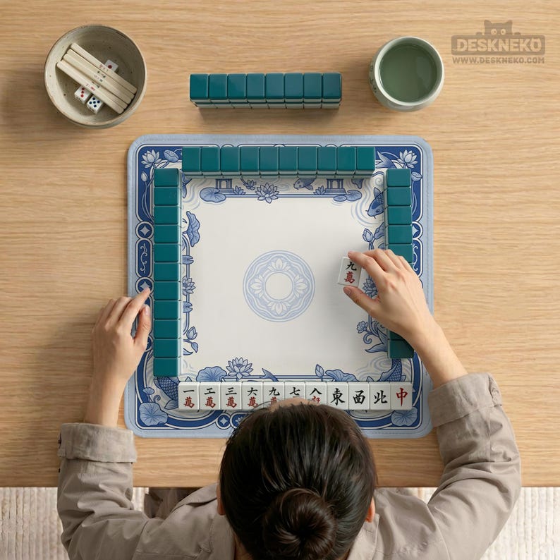 May include: A square mahjong mat with a blue and white floral border featuring lotus flowers and fish, surrounding a central circular pattern. Dark teal mahjong tiles are arranged in a square formation on the mat, with additional tiles and dice.