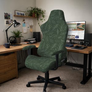 May include: A gaming chair featuring a dark green fabric cover with a white line-art pattern of leaves and stems. The chair has a high backrest, adjustable armrests, and a black five-point base with casters for mobility in a home office.